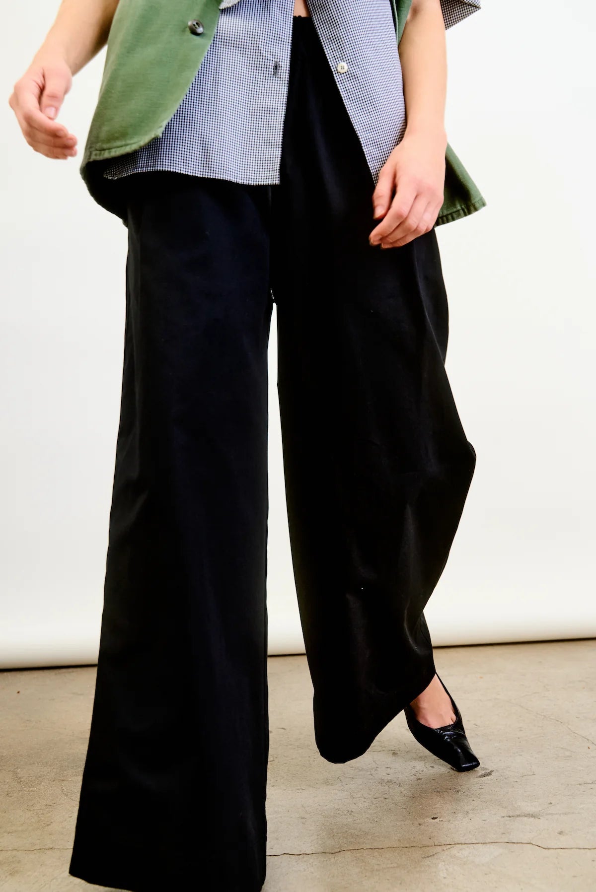 A person wears Connor Wide Leg Pant by Aquarius Cocktail, styled with a striped shirt and a green blazer. Standing on concrete, one foot slightly forward in black pointed shoes, the image highlights the outfit from shoulders down.