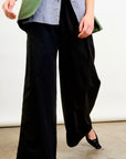 A person wears Connor Wide Leg Pant by Aquarius Cocktail, styled with a striped shirt and a green blazer. Standing on concrete, one foot slightly forward in black pointed shoes, the image highlights the outfit from shoulders down.