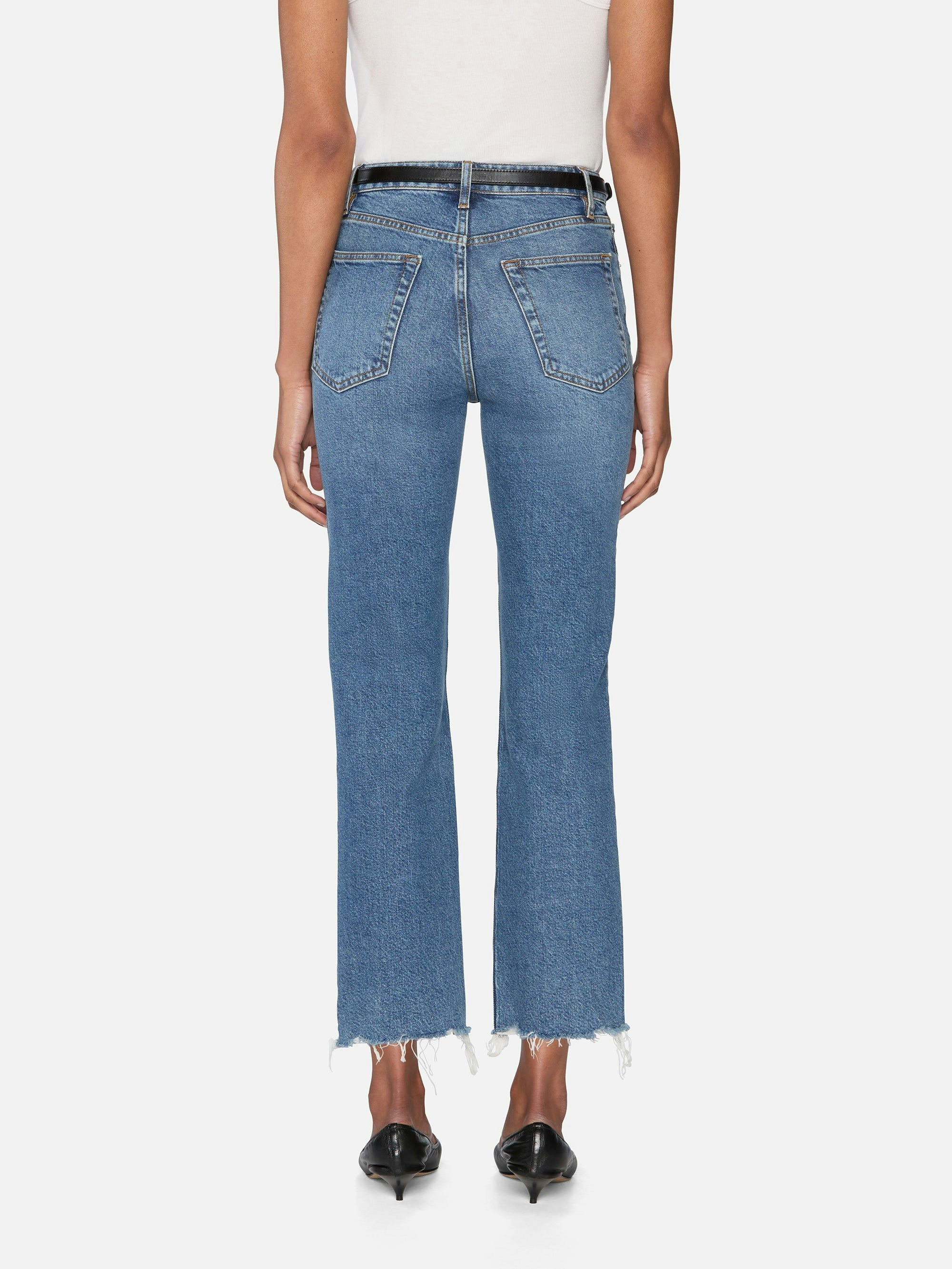 A person is seen from the back wearing FRAME’s The Ruler Crop blue high-waisted slim-straight jeans with frayed hems, two back pockets, styled with black heeled shoes and a white top.