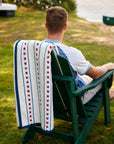 Enjoy a relaxing summer by the New England seaside with the Faire Stars & Stripes Blanket, draped over a green wooden chair outdoors near water and grass, perfect for patriotic style and comfort.