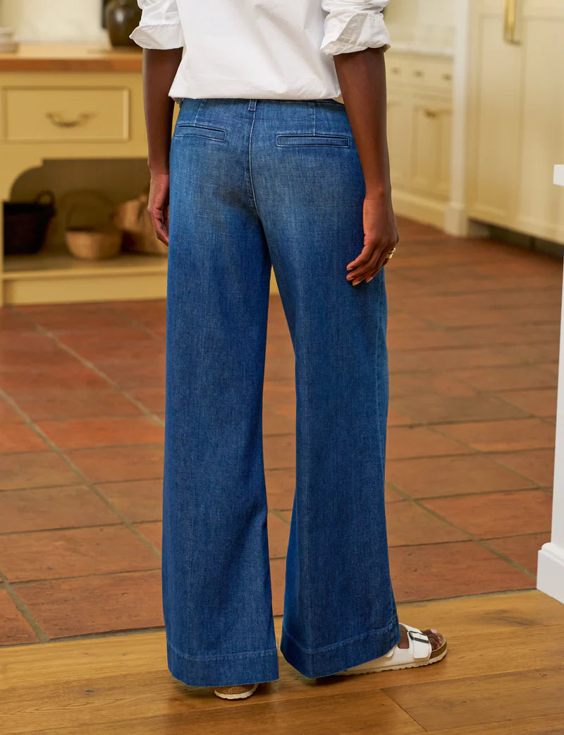 A person stands indoors on a tiled floor wearing Frank &amp; Eileen’s NYC Trouser jean in blue denim, paired with a white long-sleeve blouse and white sandals. The photo is taken from behind, showing part of a kitchen in the background.