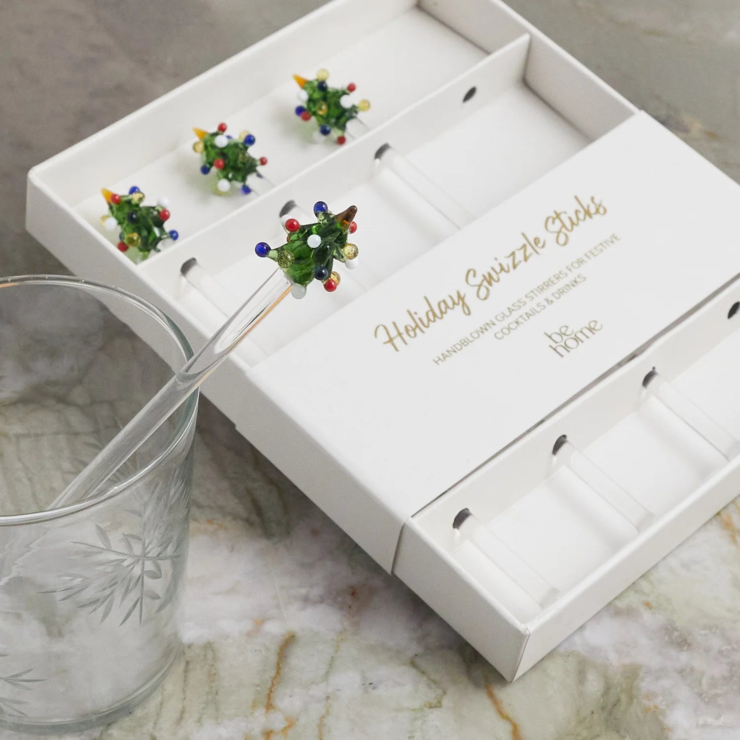 The Tree Glass Swizzle Sticks S/4 by Be Home, featuring glass tree-shaped tops with colorful dots, is displayed beside a clear glass with a frosted leaf pattern on a marble surface.