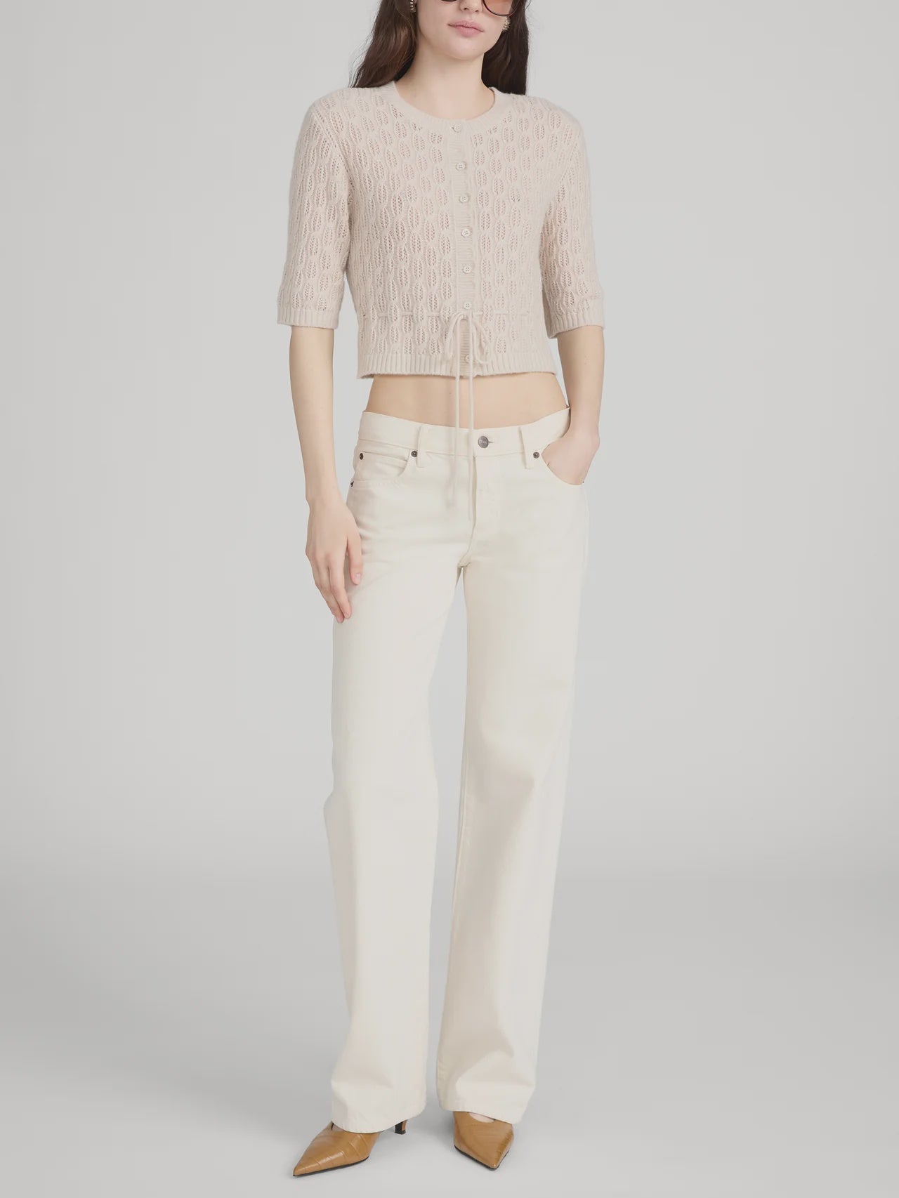 A woman stands against a plain background wearing a light pink cropped knit top, FRAME’s The Saturday Jean—100% cotton, high-waisted wide-leg jeans in off-white rigid denim—and tan pointed-toe shoes, with one hand in her pocket.