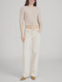 A woman stands against a plain background wearing a light pink cropped knit top, FRAME’s The Saturday Jean—100% cotton, high-waisted wide-leg jeans in off-white rigid denim—and tan pointed-toe shoes, with one hand in her pocket.