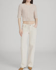 A woman stands against a plain background wearing a light pink cropped knit top, FRAME’s The Saturday Jean—100% cotton, high-waisted wide-leg jeans in off-white rigid denim—and tan pointed-toe shoes, with one hand in her pocket.