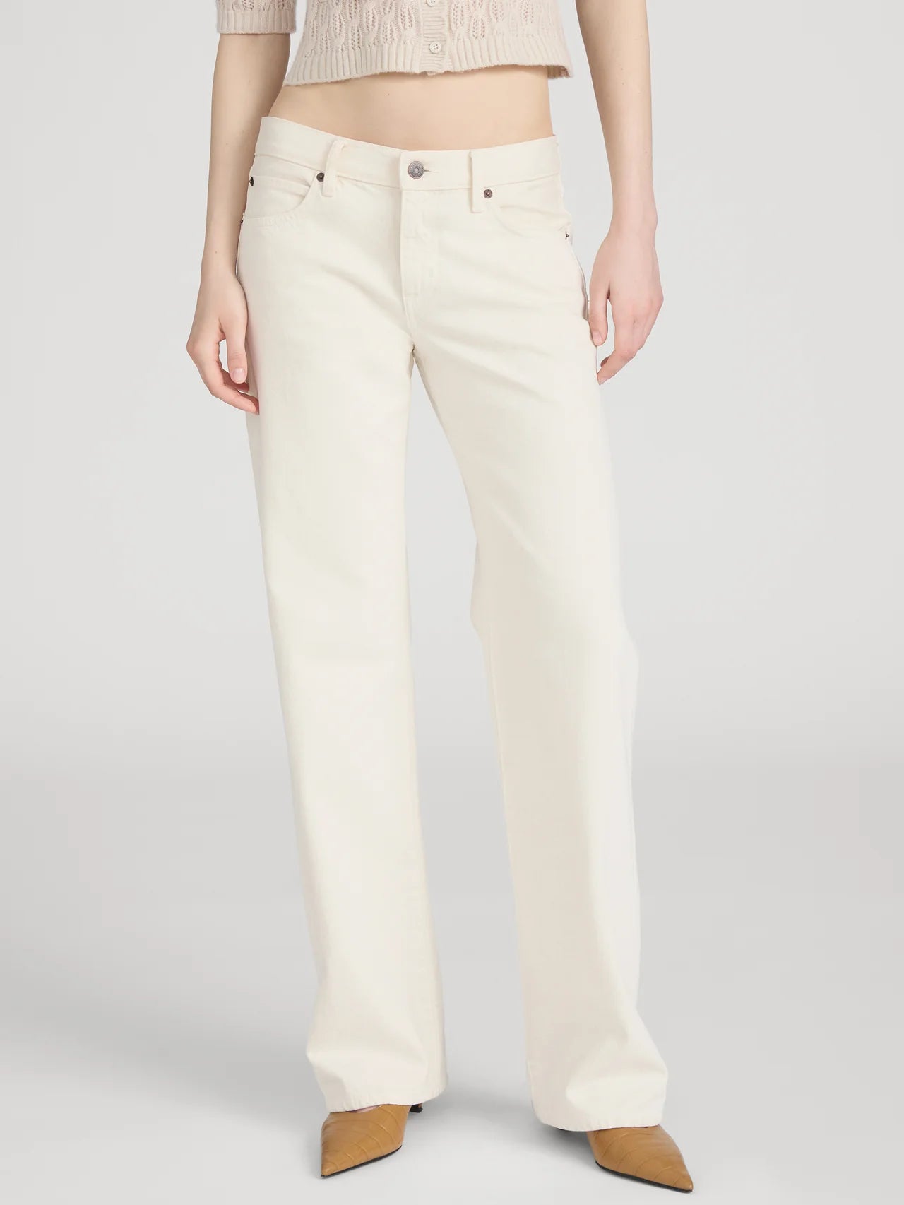 Wearing FRAME’s The Saturday Jean—high-waisted, straight-leg white jeans in 100% cotton rigid denim—with tan pointed-toe shoes and a cropped beige sweater; photo shows the look from shoulders down against a plain background.