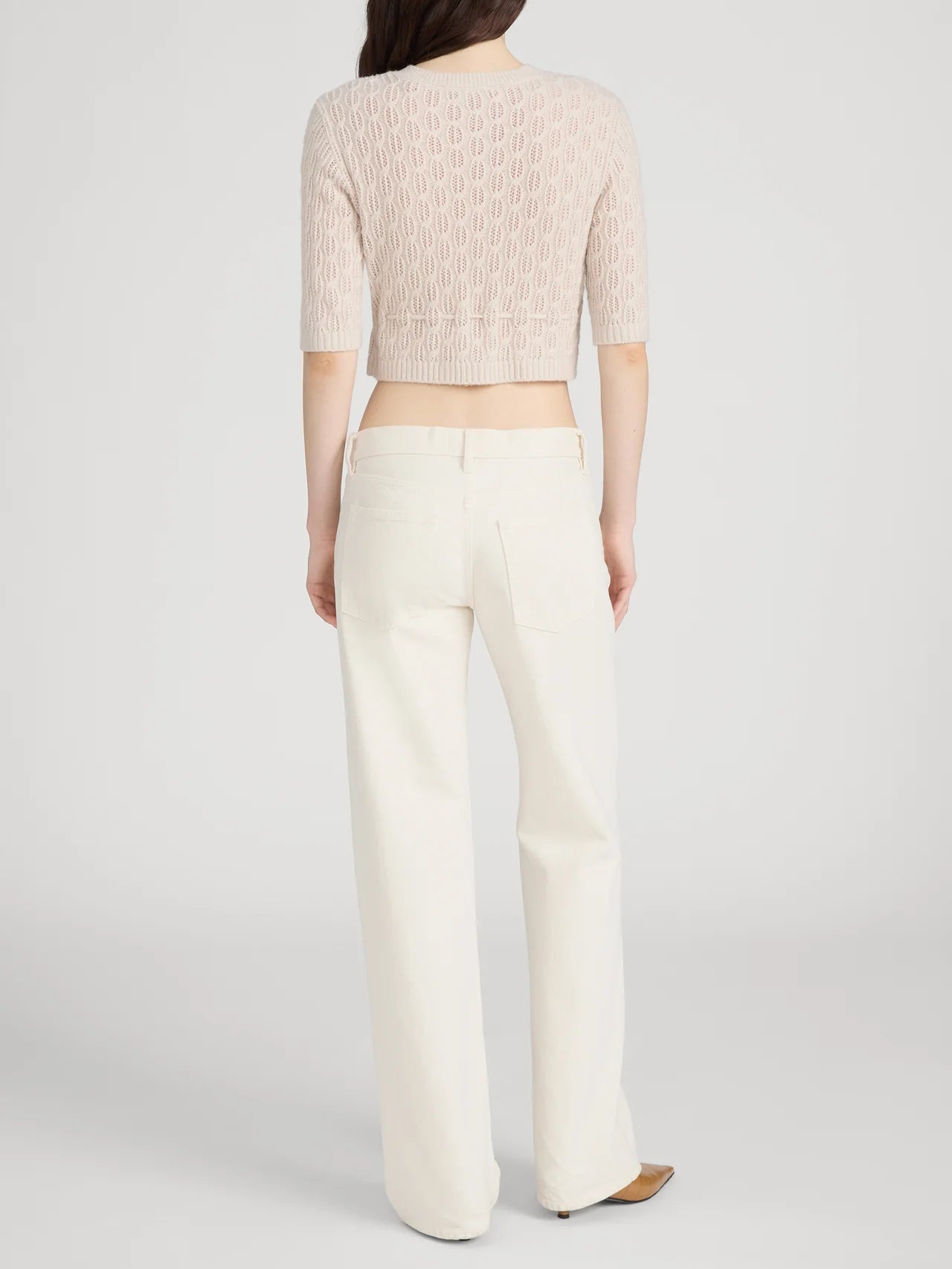 A person with long brown hair, seen from behind, wears a light pink textured knit top and FRAME’s The Saturday Jean—high-waisted, off-white 100% cotton wide-leg denim—paired with beige shoes against a plain light background.