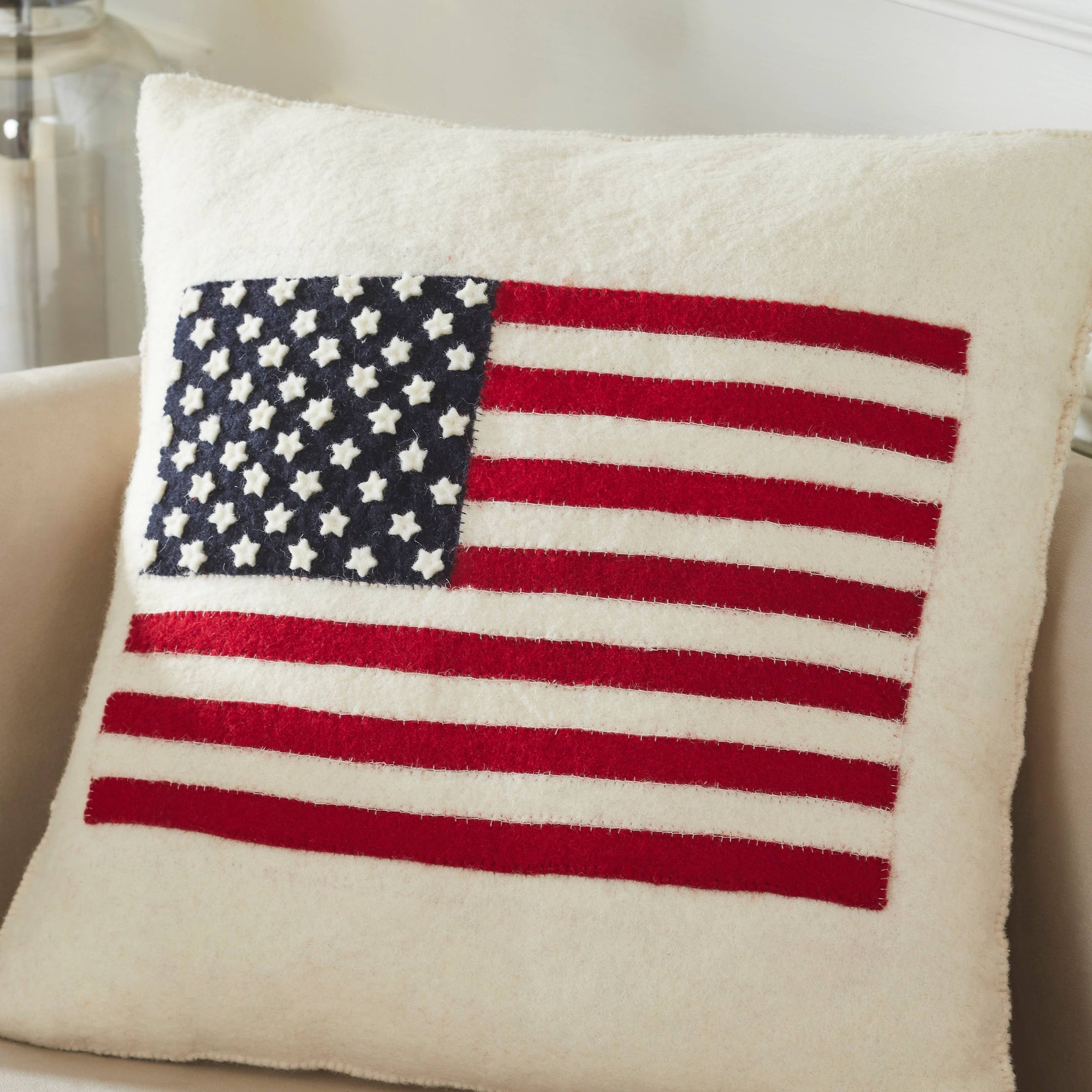 The American Flag Wool Pillow by Faire, made from hand-felted wool with blue, white, and red stitched details, sits on a beige armchair—a perfect handmade décor accent for any cozy space.