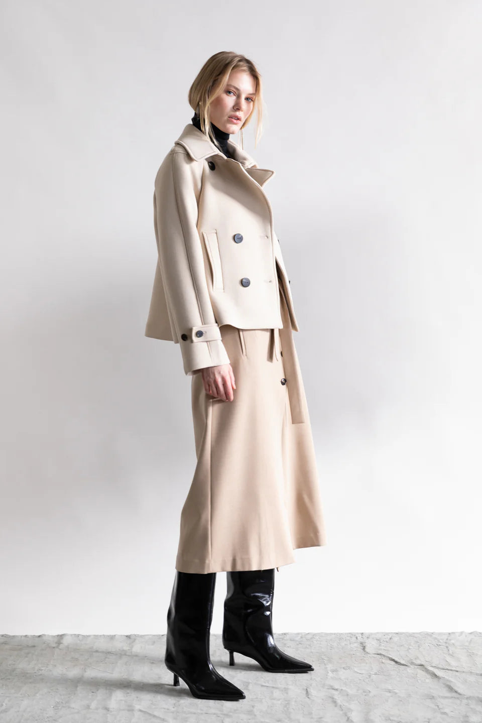 A woman models a beige midi skirt with the Psophia Cropped Blazer Jacket, posed in profile against a plain light background. She finishes the tailored look with black knee-high heeled boots and wears a neutral expression.