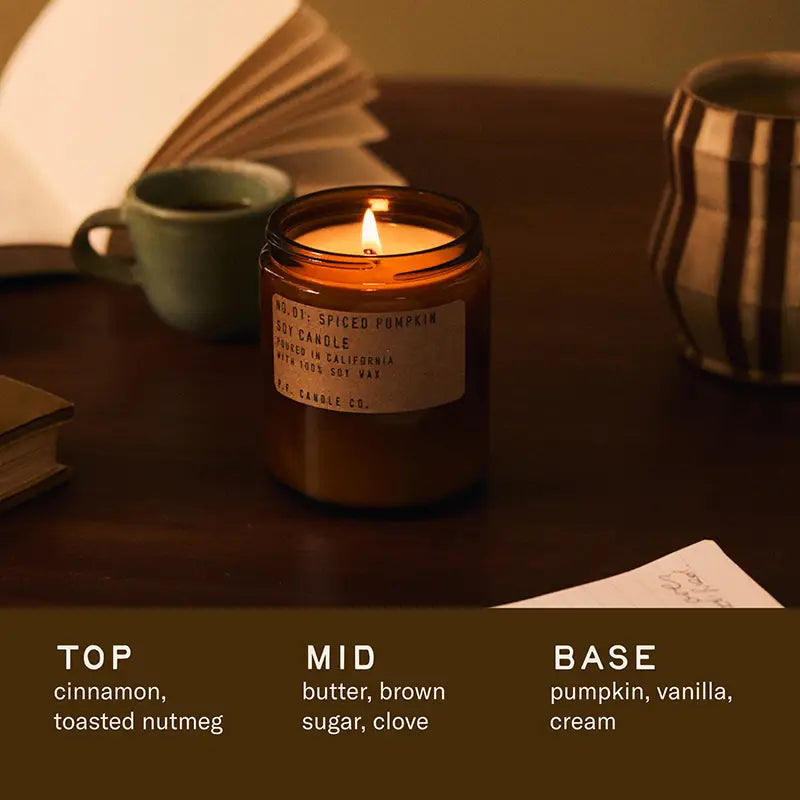 The Faire PFC Candle, a hand-poured brown glass candle, sits by books and mugs. Made with soy wax and fine fragrance oils, it features scent notes of cinnamon, toasted nutmeg, butter, brown sugar, clove, pumpkin, vanilla, and cream.