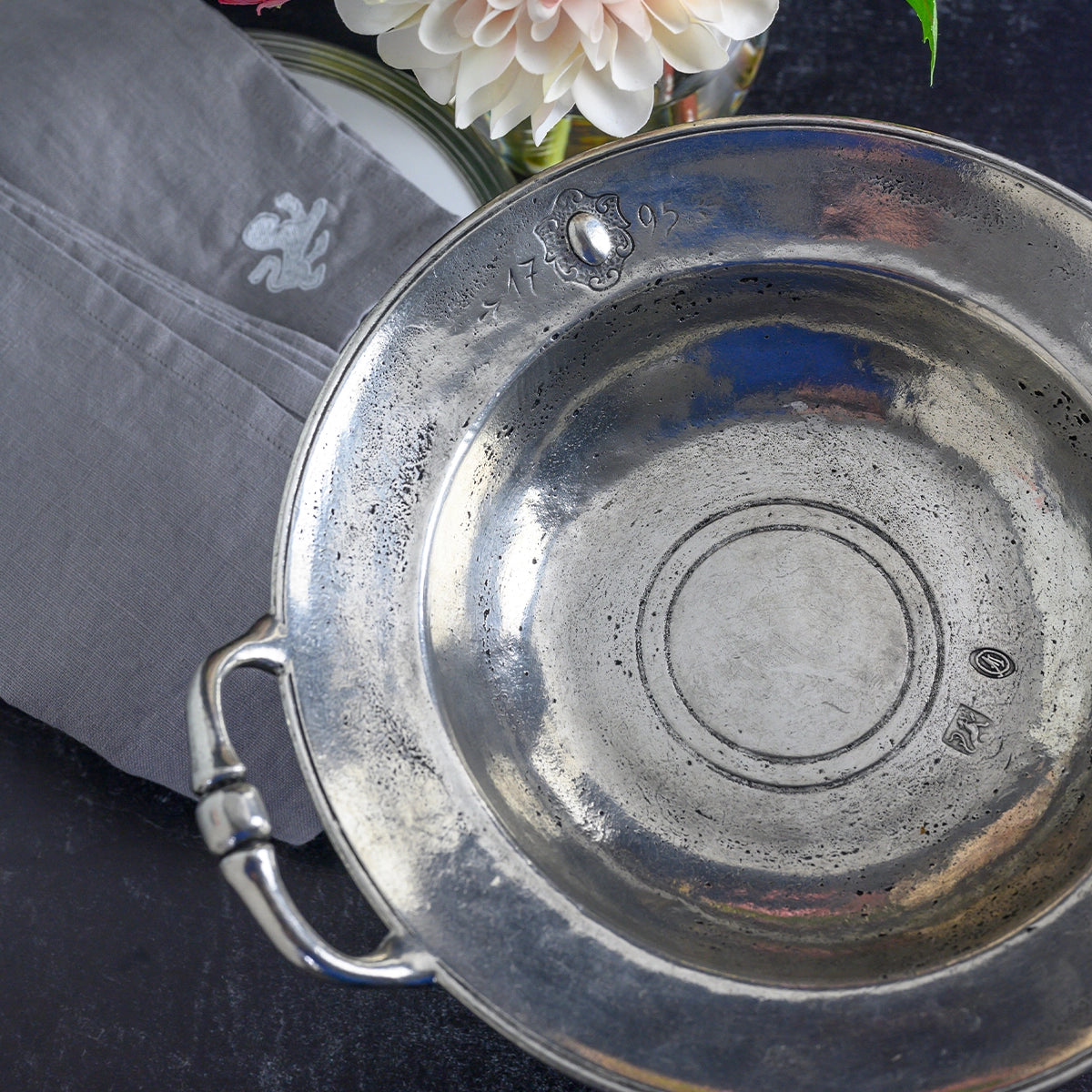 The Faire Vintage 1795 Pedestal Bowl with handles and stamped markings rests on a dark surface beside a folded embroidered gray napkin and a vase holding a white dahlia flower.