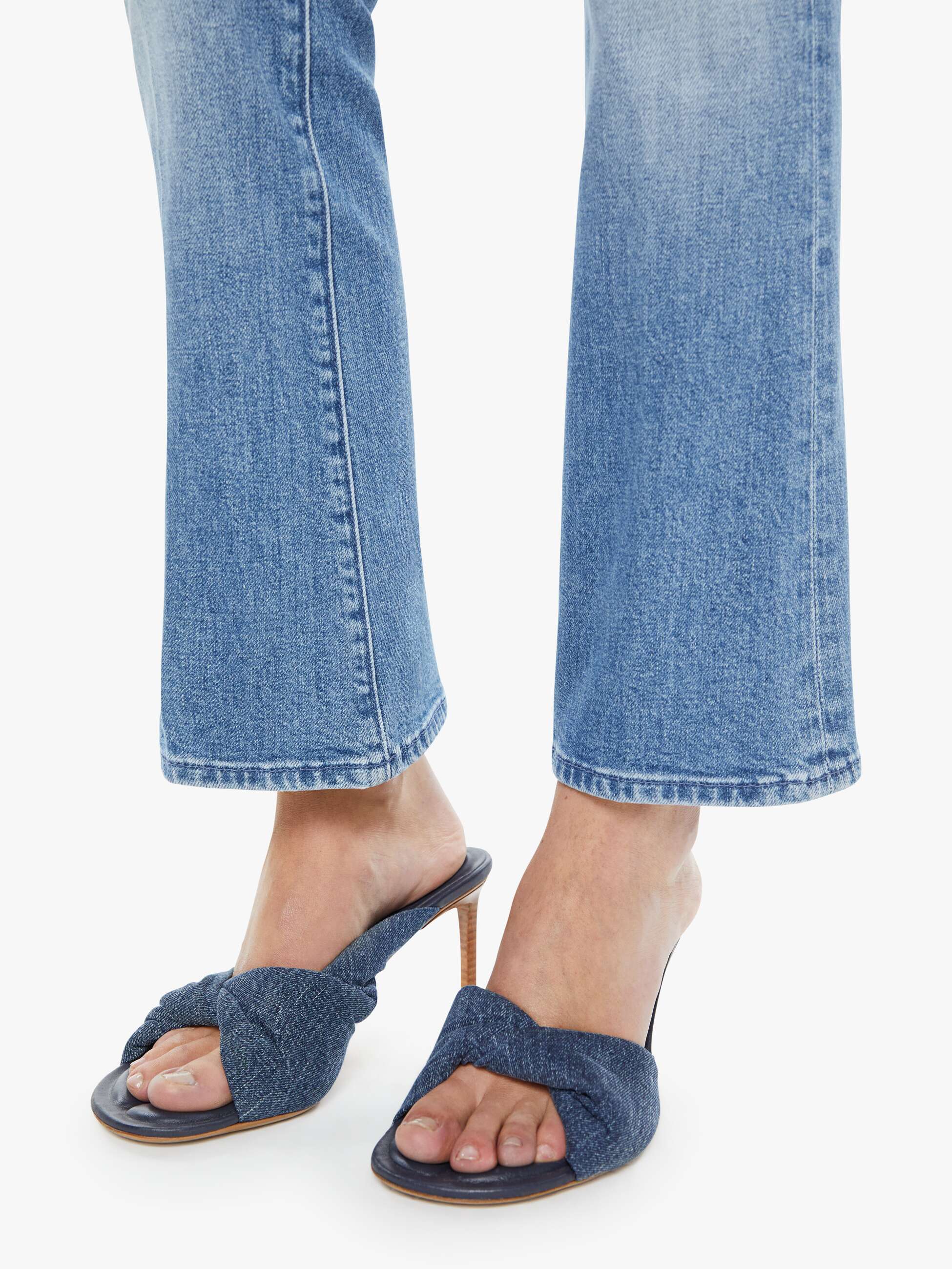 A person wearing The Outside Flood vintage blue jeans by Mother and dark blue heeled sandals with a knotted design stands on a white background.