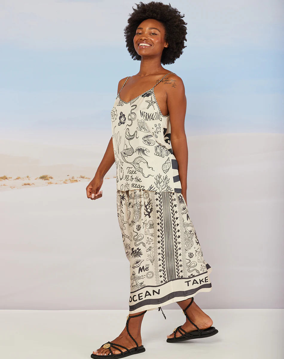 A smiling woman with short curly hair wears the Me369 Elizabeth Midi Skirt featuring black line art and text print, styled with black sandals. She stands sideways against a beach-themed backdrop for a chic summer look.