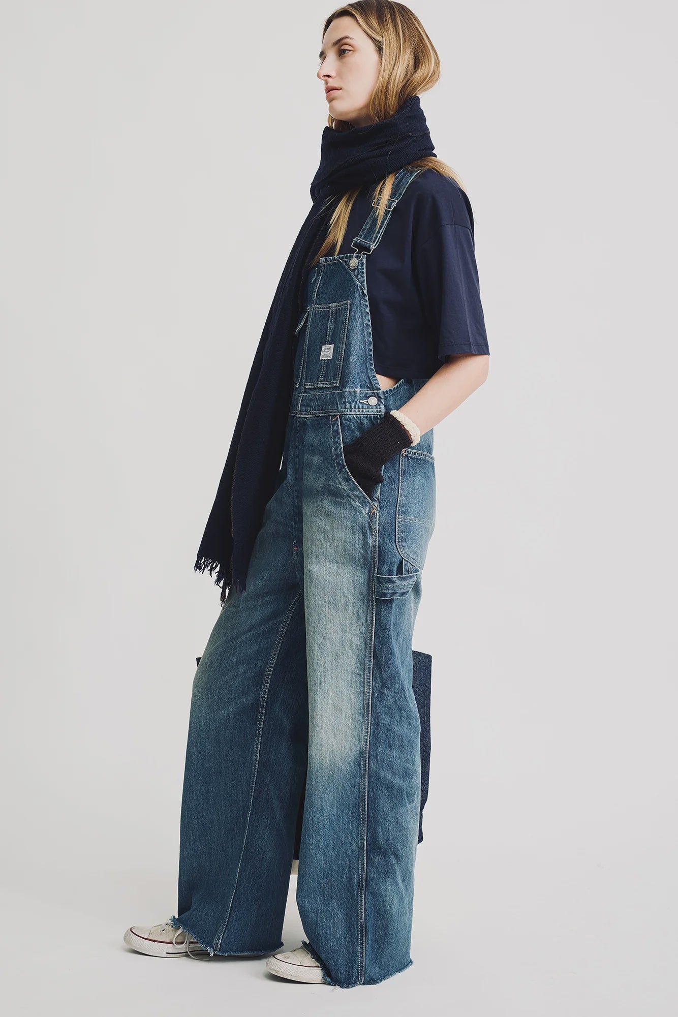 A woman stands sideways against a white background, wearing Denimist SLOUCH BIB OVERALL denim overalls, a navy t-shirt, dark scarf, fingerless gloves, and white sneakers.