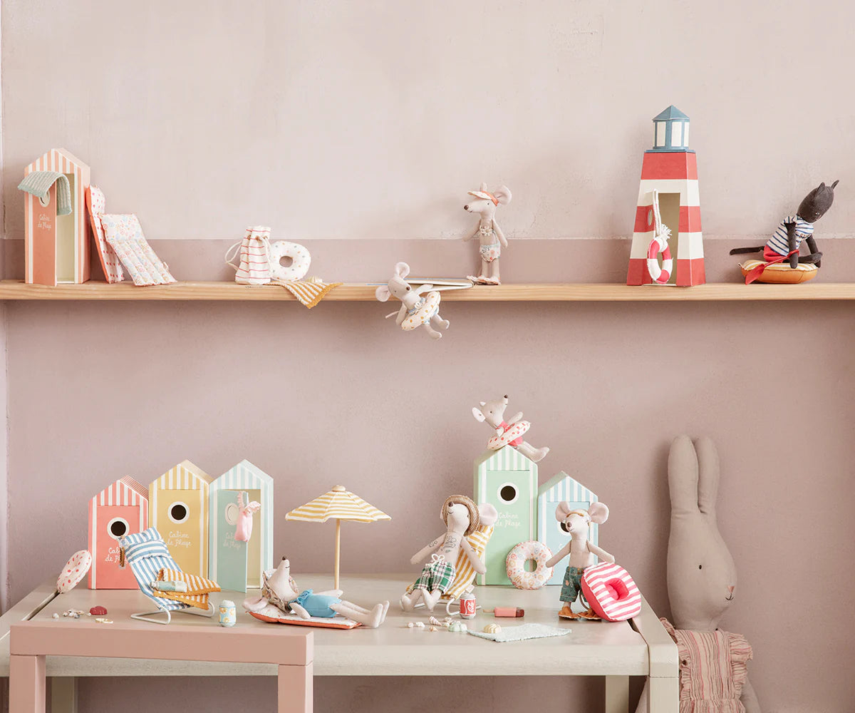 A playful Maileg display features toy mice, a rabbit, and the Bag W/ Beach Essentials set with cabins, umbrellas, striped tote, and mouse-sized accessories arranged on shelf and table against a pale pink wall.
