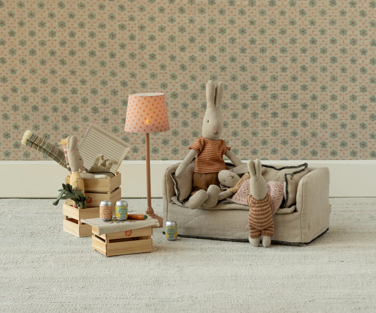 Two Maileg bunny dolls relax beside a Maileg Couch, Miniature by Maileg. Dollhouse furniture, tiny canned drinks, and a lamp create a cozy vintage room with floral wallpaper and wooden crates.