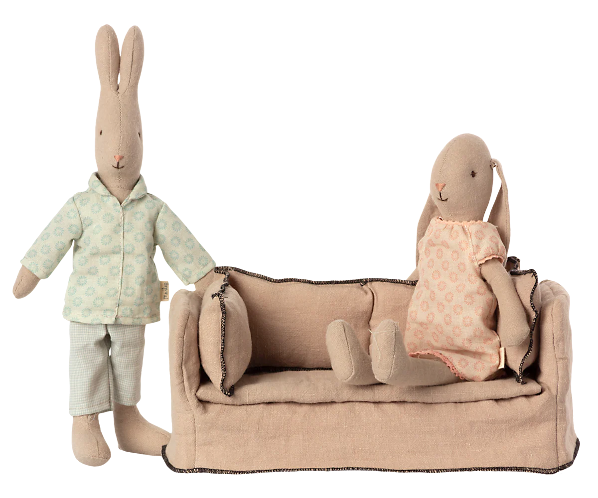 Two fabric bunny dolls sit near a Maileg Couch, Miniature by Maileg: one in pale green pajamas stands beside it, while the other in a pink dress sits on the sofa. Both are set against a plain white background.
