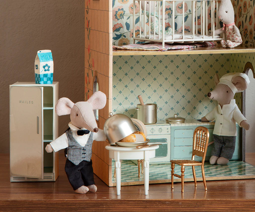 Two Maileg toy mice dressed as waiters stand in a vintage kitchen and dining room. One lifts the Maileg Cheese Bell, revealing a tasty treat, while a milk carton sits on top of the refrigerator nearby.