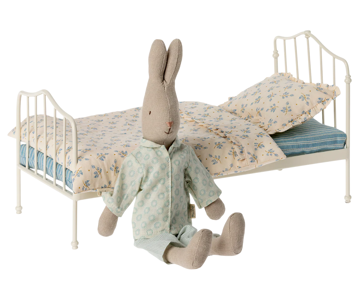 A fabric bunny doll in pajamas sits on the floor beside the Maileg Vintage Bed, Miniature by Maileg, featuring a white metal frame, floral bedding, and striped sheets in a charming vintage style.
