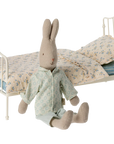 A fabric bunny doll in pajamas sits on the floor beside the Maileg Vintage Bed, Miniature by Maileg, featuring a white metal frame, floral bedding, and striped sheets in a charming vintage style.