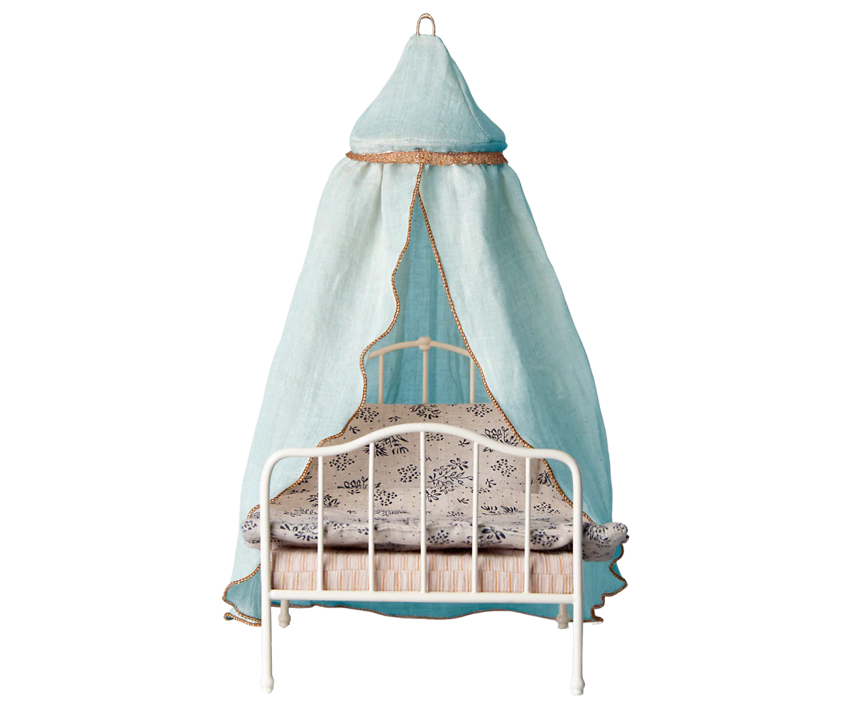 The Maileg Canopy Bed by Maileg features a small white metal frame with patterned bedding and a light blue fabric canopy trimmed in gold, draped from an overhead ring—a charming touch for any bedroom decor.