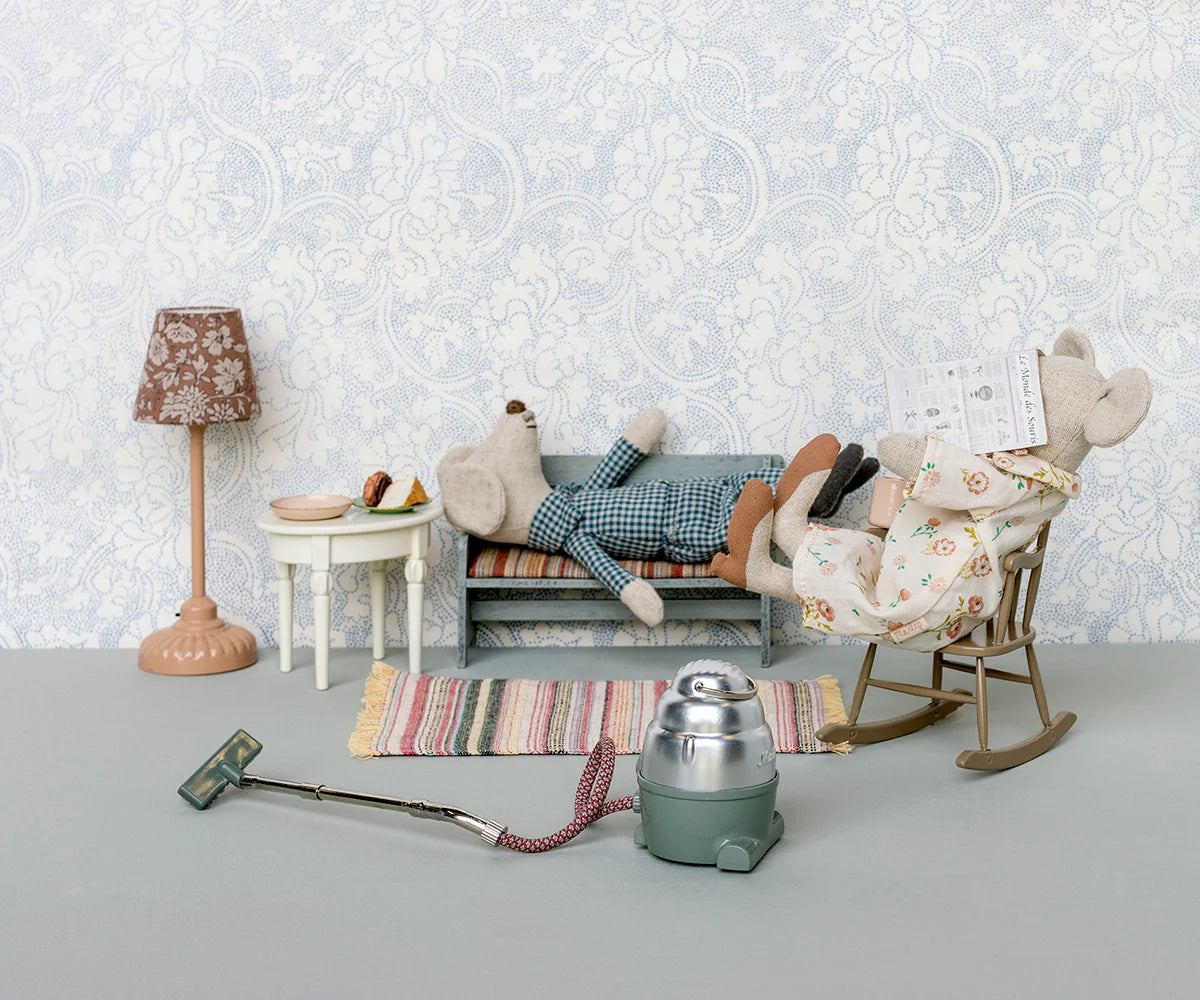 Two Maileg mouse dolls lounge in a tiny living room with Maileg furniture, including the Maileg Bench. One mouse reclines, another reads in a rocking chair, with a food table, lamp, vacuum, and rug completing the charming scene.