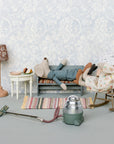 Two Maileg mouse dolls lounge in a tiny living room with Maileg furniture, including the Maileg Bench. One mouse reclines, another reads in a rocking chair, with a food table, lamp, vacuum, and rug completing the charming scene.