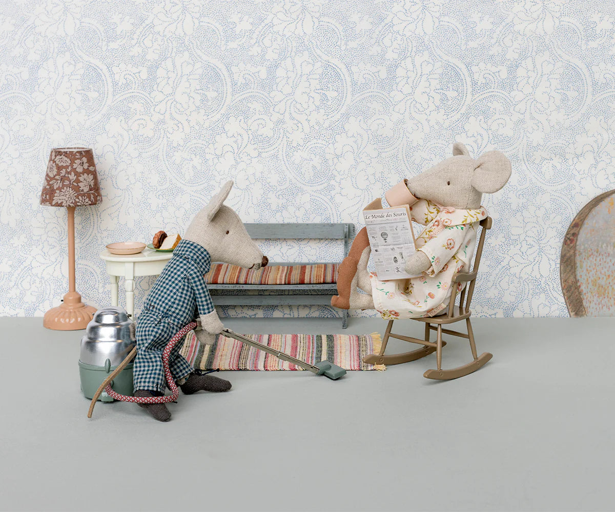 Two Maileg toy mice in a vintage room: one reads the paper in a rocking chair, while the other, in a checkered robe, cleans the floor with the Maileg Vacuum Cleaner. A striped bench and lamp complete the scene.