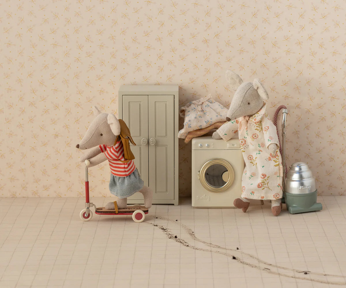 Two Maileg toy mice in a dollhouse scene: one rides a red scooter, the other vacuums by a washing machine. Tiny clothes, furniture, and the Maileg Closet appear in the background. Scooter tracks are visible on the floor.