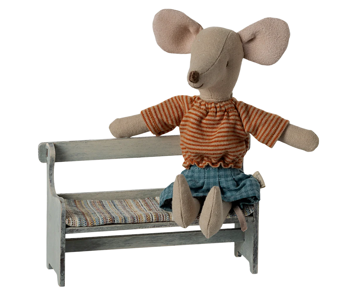 A Maileg stuffed mouse in a striped orange shirt and denim shorts sits on the Maileg Bench, a charming piece featuring a striped cushion. The mouse’s big ears and stitched nose add to this adorable Maileg scene.