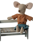 A Maileg stuffed mouse in a striped orange shirt and denim shorts sits on the Maileg Bench, a charming piece featuring a striped cushion. The mouse’s big ears and stitched nose add to this adorable Maileg scene.