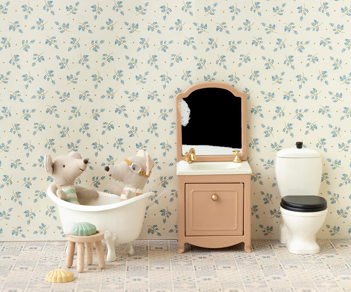 Two Maileg toy mice sit in a white bathtub in a dollhouse bathroom with a vanity, mirror, and the Maileg Toilet by Maileg. Light floral wallpaper and tiled flooring enhance the cozy, whimsical scene.