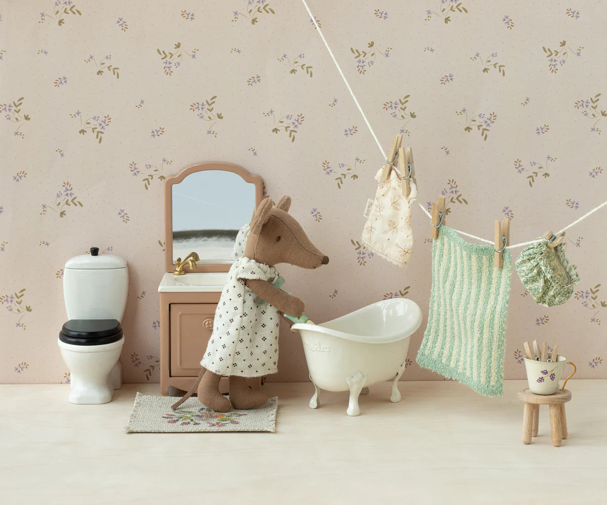 A Maileg Sink Dresser stands in a miniature bathroom scene with a toy mouse in a dress, white bathtub, toilet, mirror, and laundry on a clothesline against charming floral wallpaper.