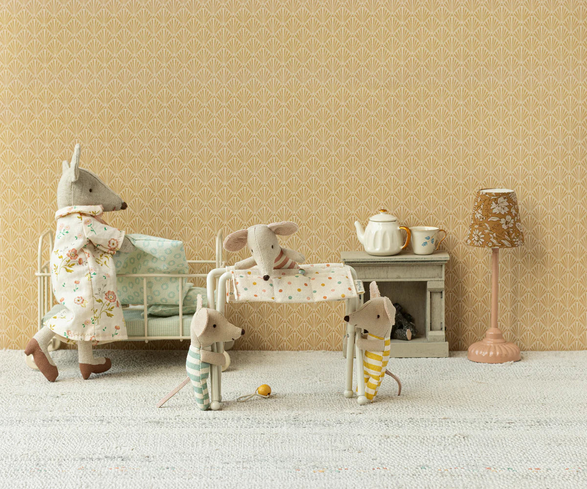Four Maileg toy mice in a vintage-style room: one adult mouse in a floral dress, two small mice in stripes, and a baby mouse asleep in a crib. The Maileg Nursery Table holds baby accessories beside the bed, with lamp, teapot set, and patterned wallpaper.