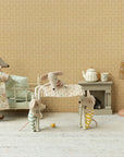 Four Maileg toy mice in a vintage-style room: one adult mouse in a floral dress, two small mice in stripes, and a baby mouse asleep in a crib. The Maileg Nursery Table holds baby accessories beside the bed, with lamp, teapot set, and patterned wallpaper.