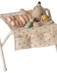A Maileg Nursery Table by Maileg features a small plush baby mouse in striped pajamas, lounging with a yellow pacifier and green bottle, surrounded by charming baby accessories.