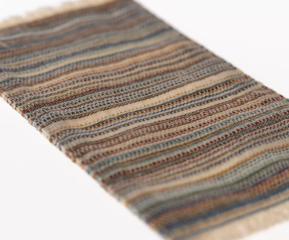 A close-up of the Maileg Rug by Maileg shows its woven texture with horizontal stripes in earthy browns, blues, and beige on a white background—perfect as a distinctive door mat or small decor item.
