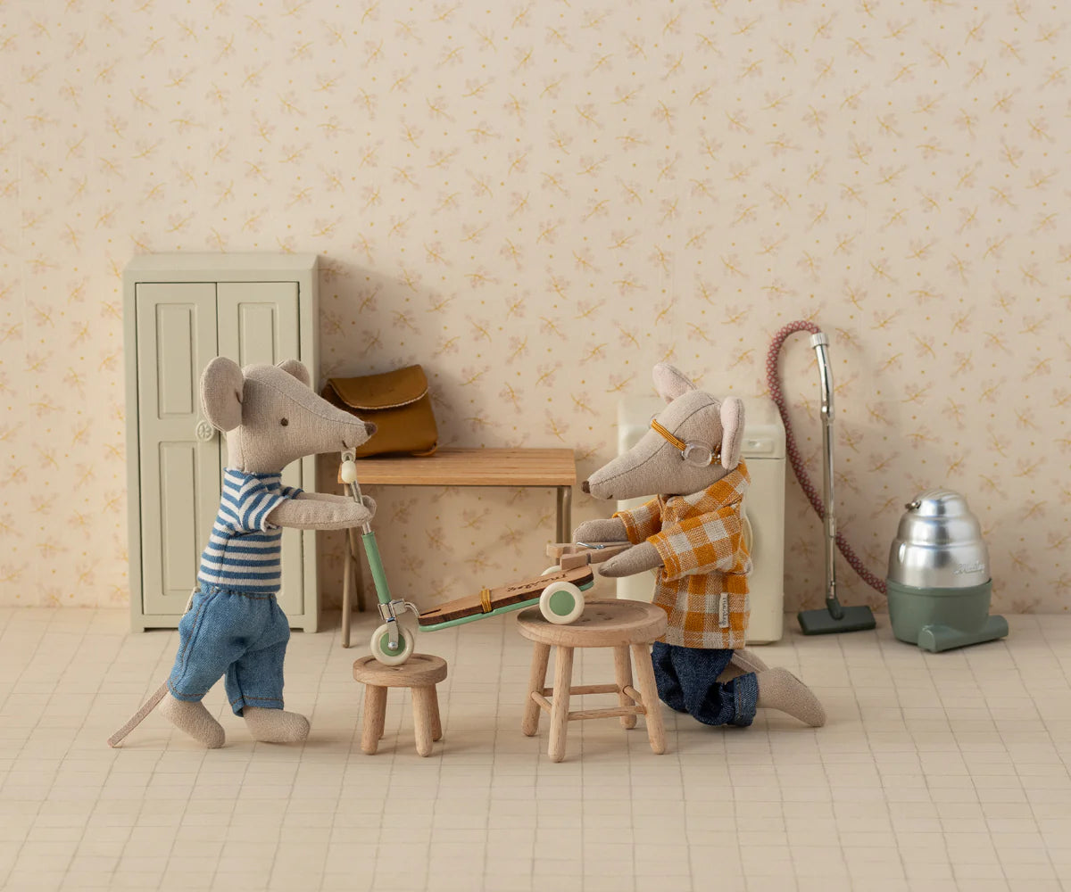 Two Maileg mice with magnets in their paws play indoors with the Maileg Kick Board. One wears a striped shirt and jeans, the other kneels in plaid. Miniature Maileg furniture and a tiny vacuum decorate the scene.