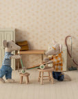 Two Maileg mice with magnets in their paws play indoors with the Maileg Kick Board. One wears a striped shirt and jeans, the other kneels in plaid. Miniature Maileg furniture and a tiny vacuum decorate the scene.