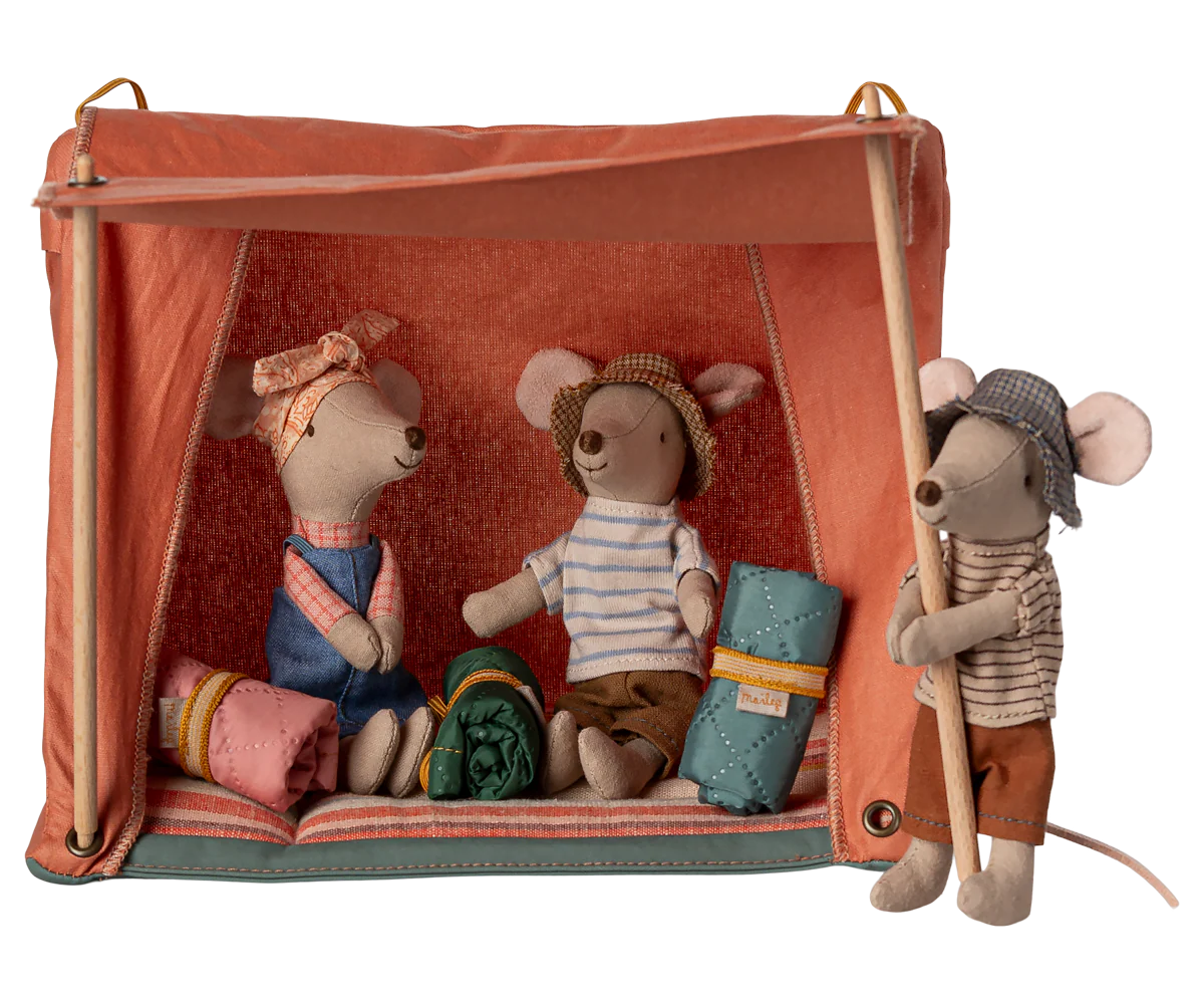 Three Maileg mice prepare for adventure with the Maileg Happy Camper Mouse Tent—two nap in sleeping bags inside the bright orange tent, while another stands outside with a wooden pole, all ready for imaginative play.