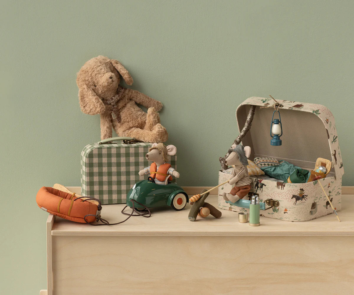 A Maileg Rubber Boat by Maileg, complete with a life vest, is displayed on a light wood dresser with hiker mice, camping gear like a tent and lantern, a soft toy dog on a plaid suitcase, and a toy car—all against a pale green wall.