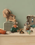 A Maileg Rubber Boat by Maileg, complete with a life vest, is displayed on a light wood dresser with hiker mice, camping gear like a tent and lantern, a soft toy dog on a plaid suitcase, and a toy car—all against a pale green wall.
