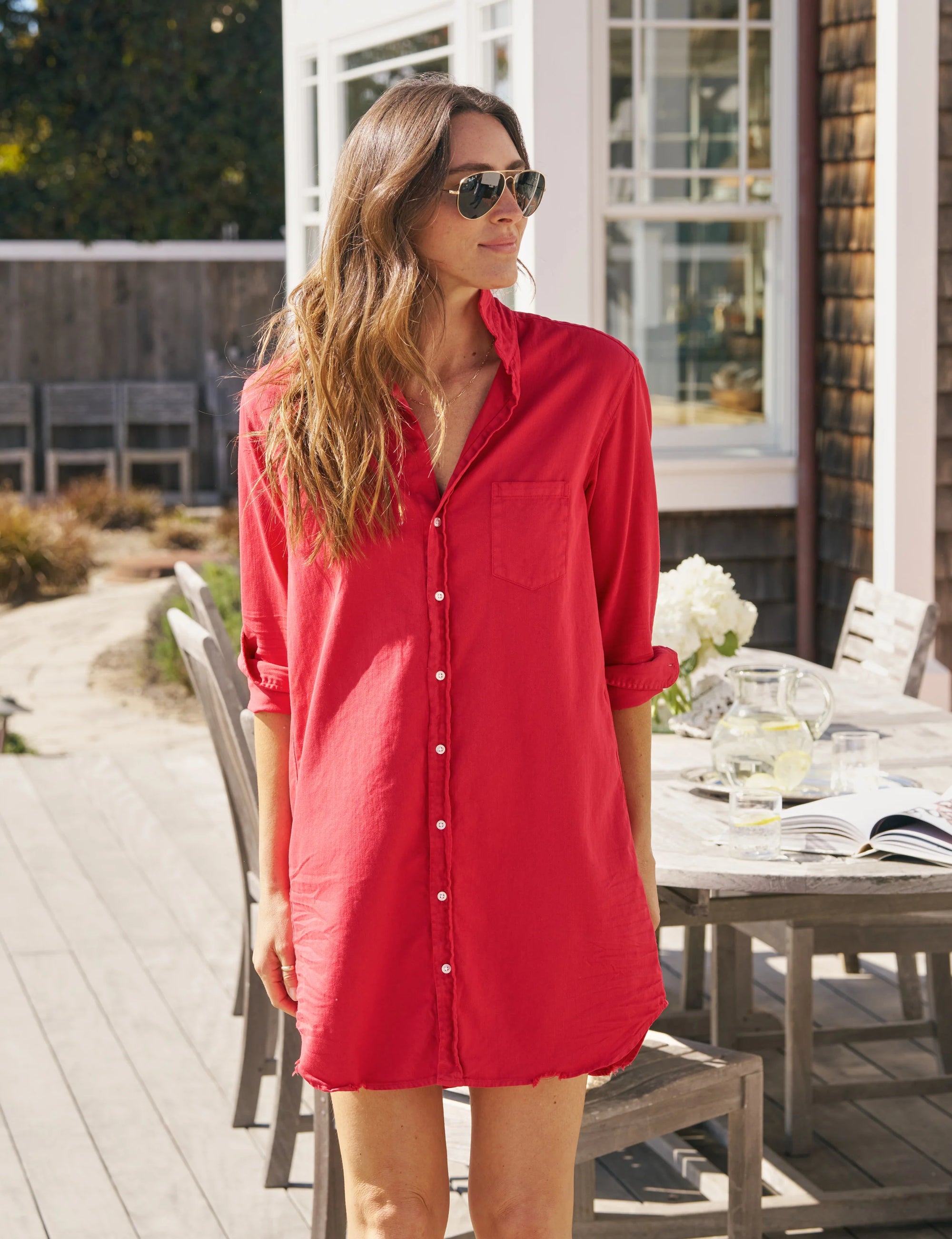 A woman in sunglasses and the Frank &amp; Eileen MARY Shirtdress S25 stands outdoors by a wooden patio table with chairs, books, and glasses, enjoying the sunlight.