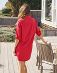 A woman with long brown hair wears the Frank & Eileen MARY Shirtdress S25 in bright red, styled with rolled-up sleeves and button closure, standing on a wooden deck outdoors and resting her hand on a chair.