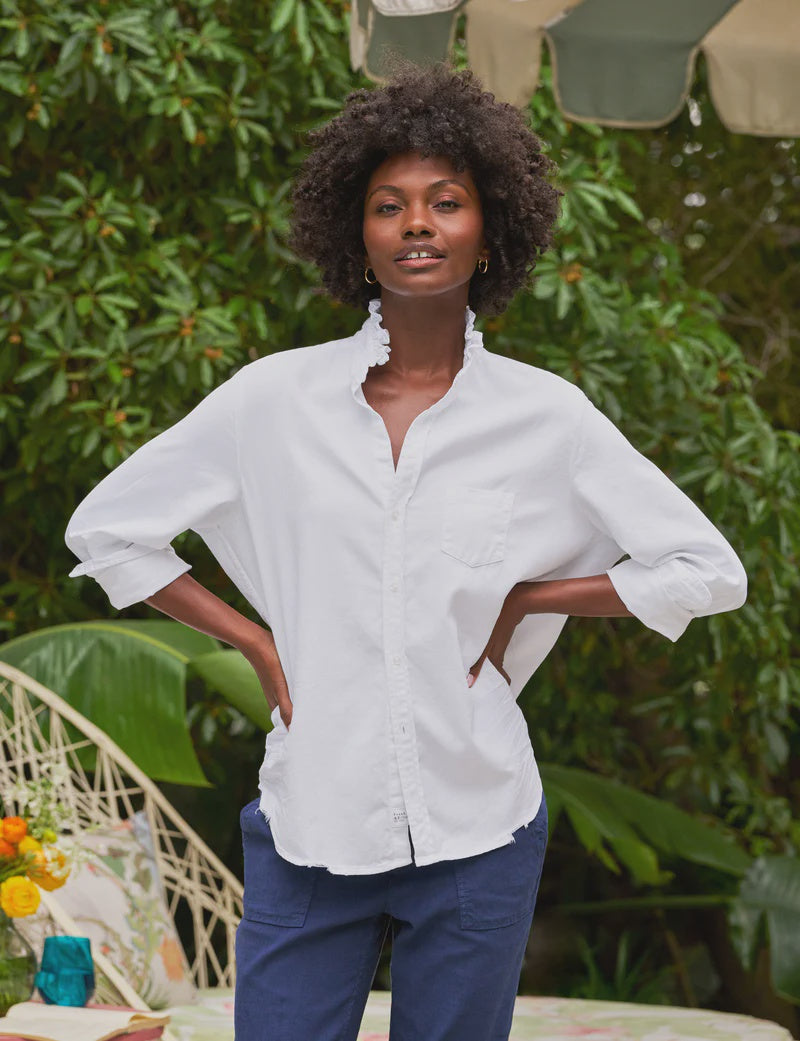 A person with curly hair stands outdoors, hands on hips, wearing the Frank & Eileen Ruffle/Eileen Button Up in white and blue pants. Green foliage and patio furniture can be seen in the background.
