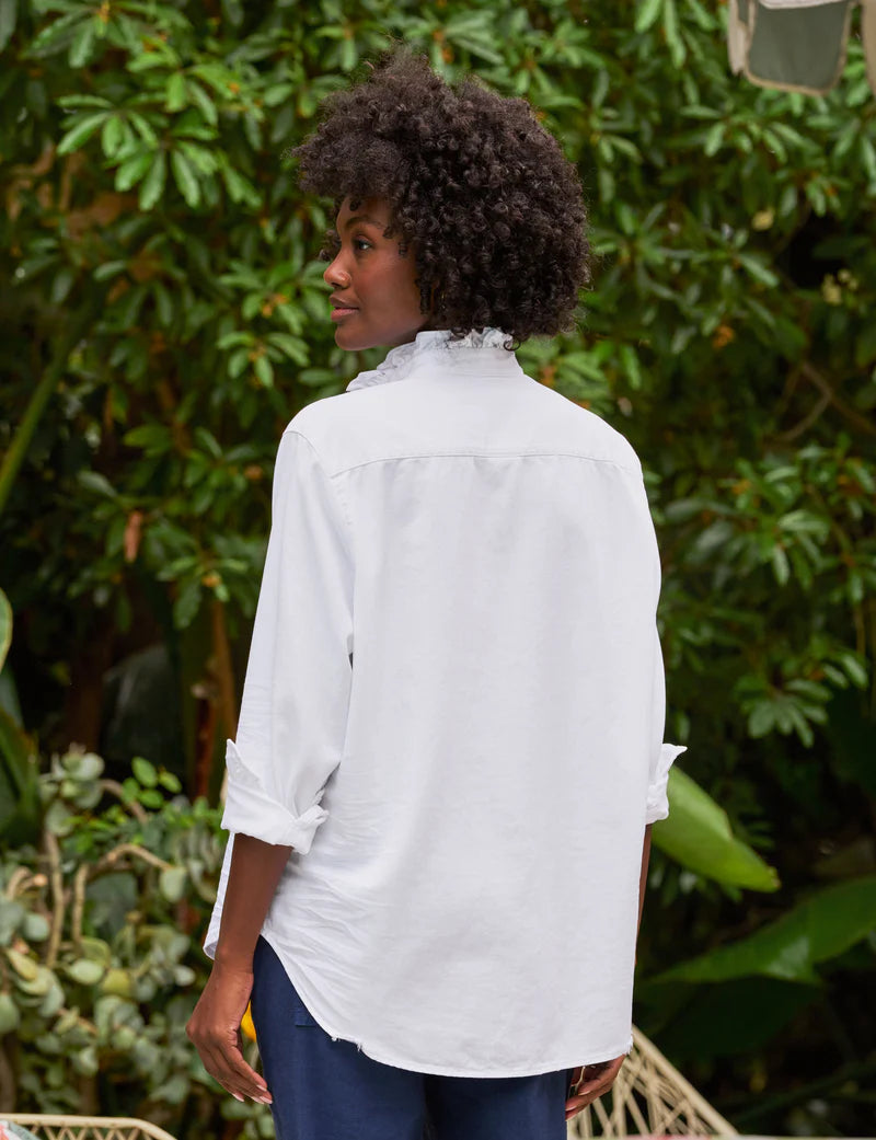 A person with curly hair stands outdoors, facing away, wearing the Frank & Eileen Ruffle/Eileen Button Up with rolled-up sleeves. Green leafy plants fill the background.