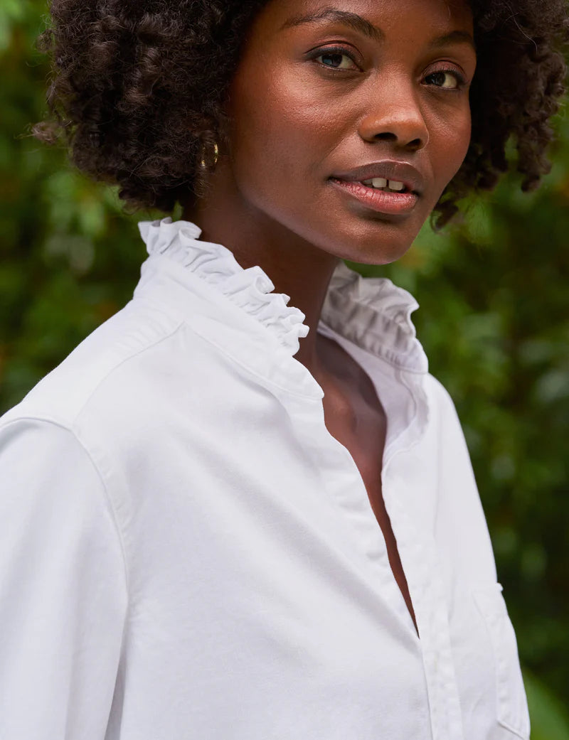 A woman with short curly hair wears the Frank & Eileen Ruffle/Eileen Button Up, a flattering white shirt with a ruffled collar, standing outdoors in front of green foliage.
