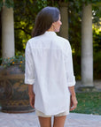 A woman with long brown hair stands outdoors in a garden, wearing the Frank & Eileen Victoria Collarless tuxedo shirt with a woven jacquard bib and rolled-up sleeves, paired with light shorts.
