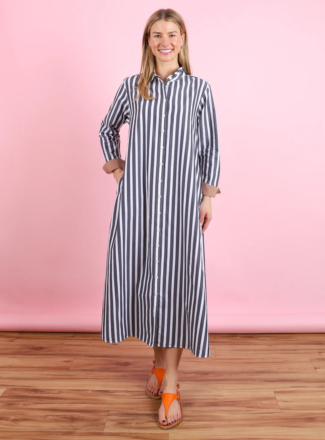 A smiling woman stands on a wooden floor with a pink background, wearing the Xirena Boden Dress, a striped cotton poplin shirtdress with front buttons, paired with orange sandals.