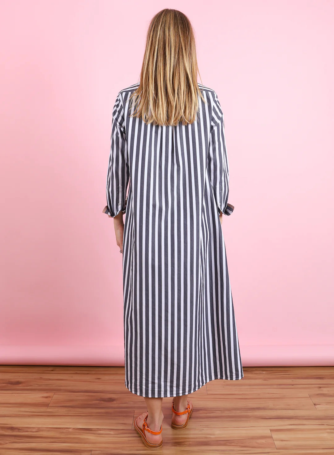 A woman with long blonde hair faces away wearing the Xirena Boden Dress—an ankle-length, loose-fit, long-sleeve cotton poplin shirtdress with vertical black and white stripes—and orange sandals, against a pink background and wooden floor.